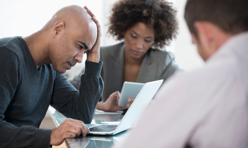 Man at a table with colleagues, appearing frustrated while working on ASD Essential Eight cybersecurity strategies.
